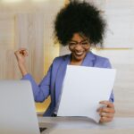 A cheerful businesswoman in a purple blazer smiles and raises her fist in excitement while looking at a document at her desk with a laptop.