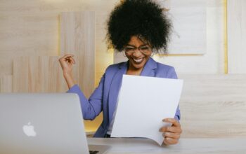 A cheerful businesswoman in a purple blazer smiles and raises her fist in excitement while looking at a document at her desk with a laptop.