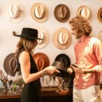 Woman wearing black hat choosing another hat while talking to a man in a stylish hat shop with many hats displayed on the wall.