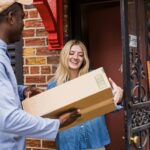 A smiling woman receives a package delivery at her doorstep, representing understanding customer satisfaction and motivation.